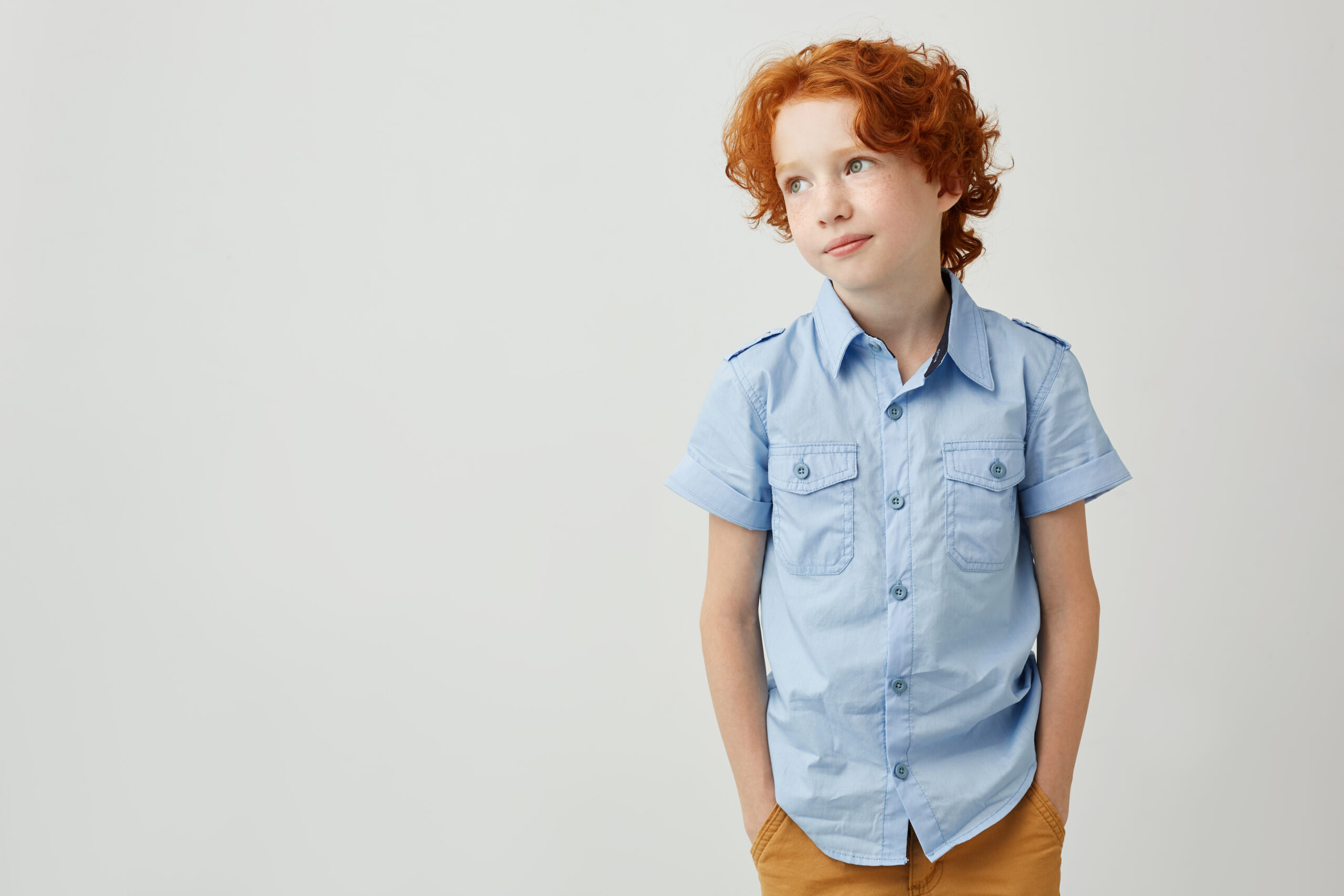 Portrait o little ginger boy with freckles holding hands in pockets, gently smiling and looking aside on white background. Copy space.